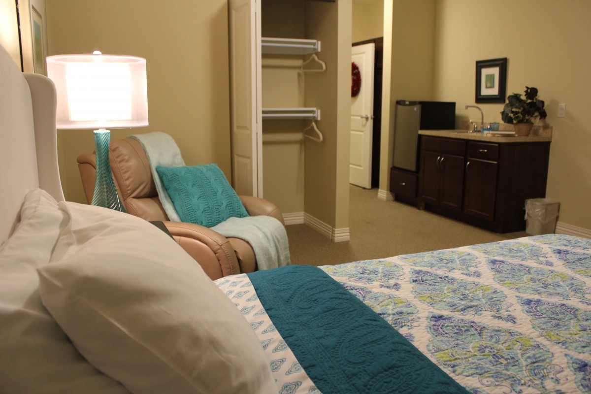 A cozy bedroom featuring a neatly made bed with blue and white bedding, an armchair, and a small kitchenette in the background.
