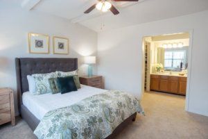 A cozy bedroom with a neatly made bed, featuring decorative pillows and a floral comforter. A ceiling fan hangs above, and artwork adorns the wall. A lamp sits on the bedside table, and the room connects to an adjacent bathroom.