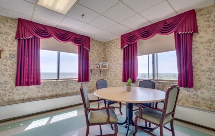 A small conference room with a round table, surrounded by chairs and large windows with pink curtains.