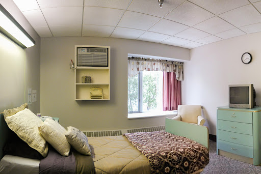 A comfortable hospital room featuring a bed, an air conditioning unit, and a cozy sitting area by the window.
