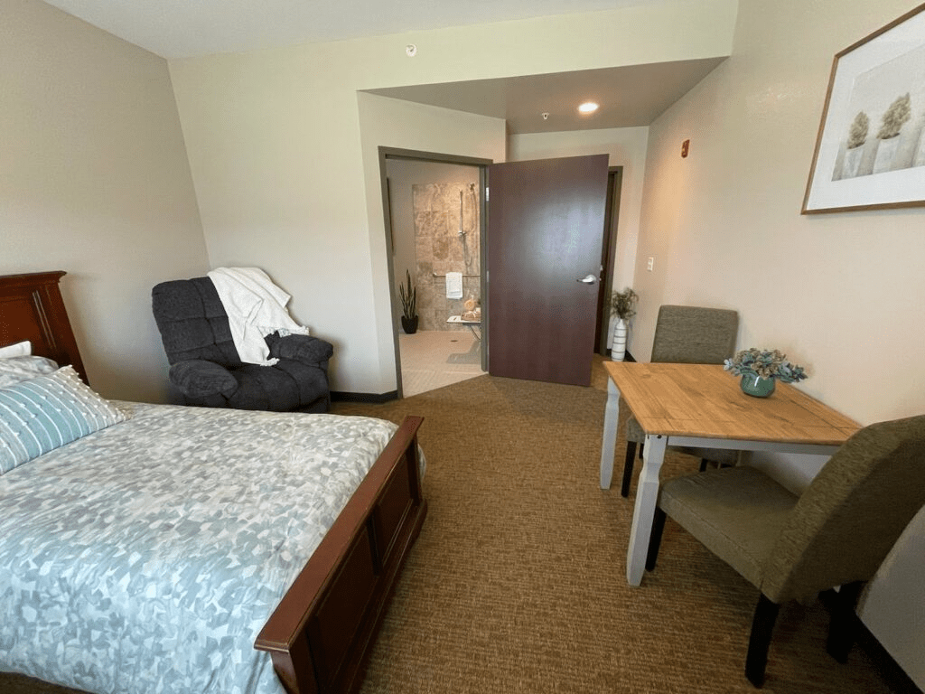 A cozy bedroom with a bed, chair, and table, leading to a bathroom.