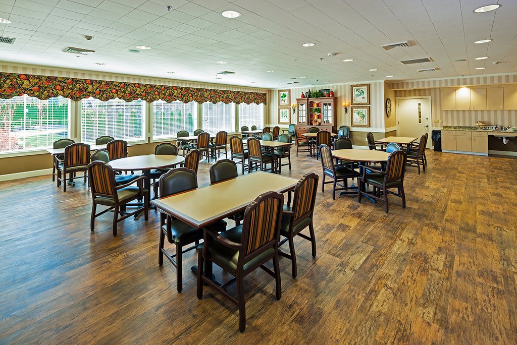 An empty dining area with multiple tables and chairs, featuring wooden flooring and large windows.