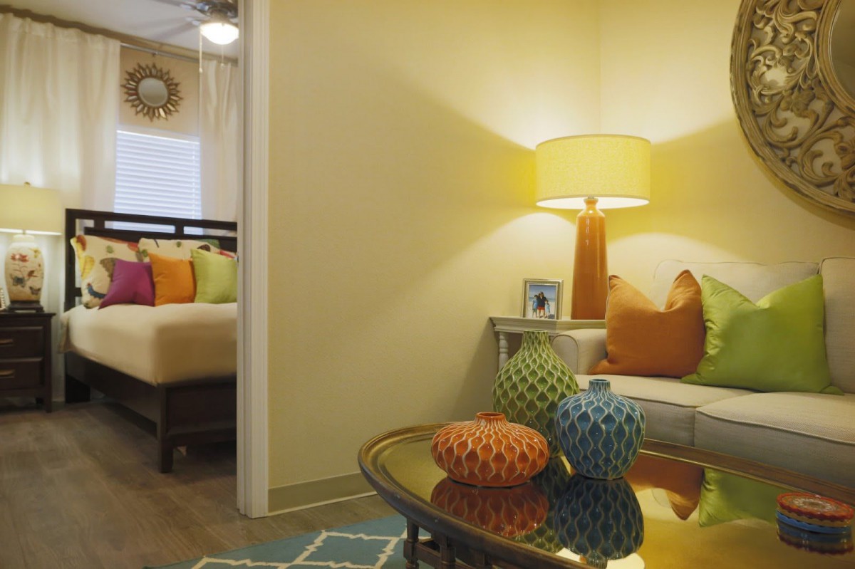 A cozy living room with colorful pillows and a stylish lamp, leading into a bedroom.