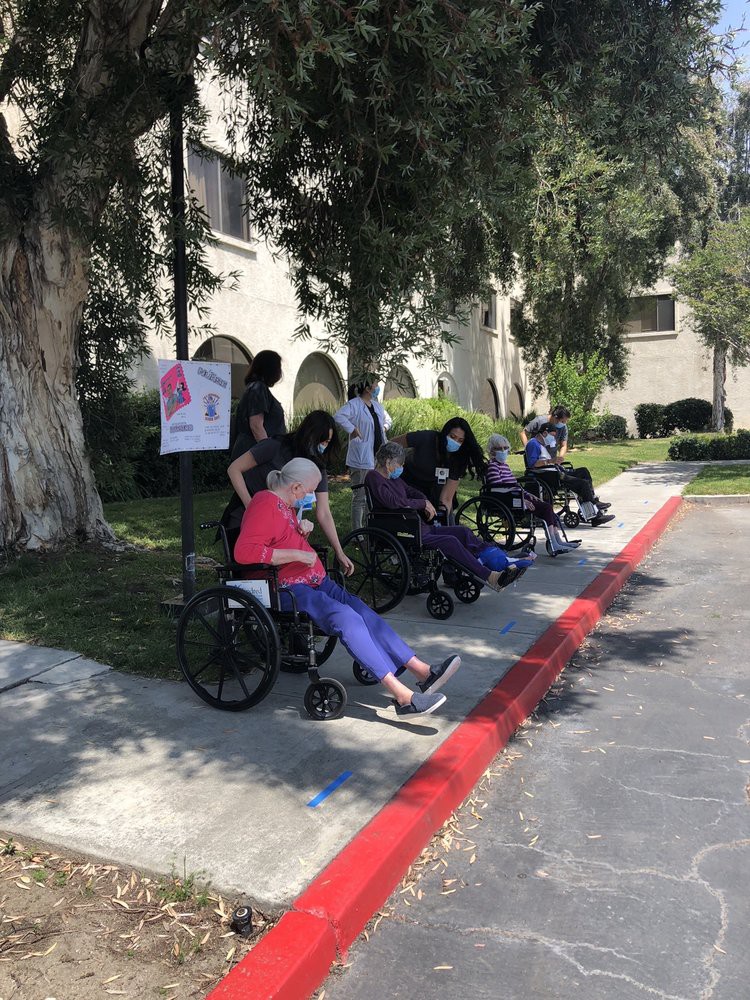 A group of elderly individuals in wheelchairs sitting on a sidewalk outside, surrounded by trees.