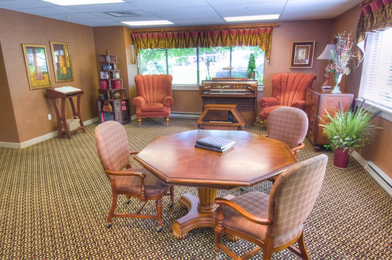 A warmly decorated conference room with a central wooden table surrounded by upholstered chairs, complete with a bookcase and large window providing natural light.