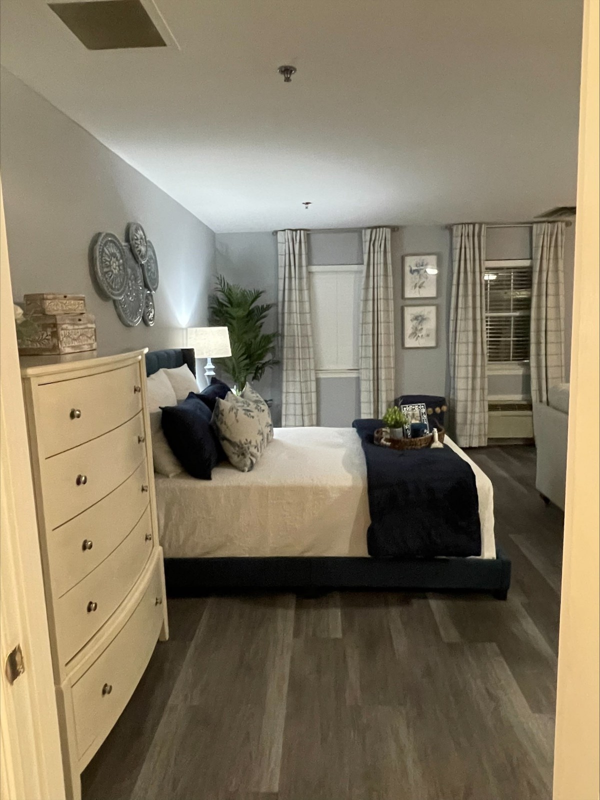 A cozy bedroom featuring a neatly made bed with navy and white bedding, a dresser on the left, and patterned curtains framing the windows.