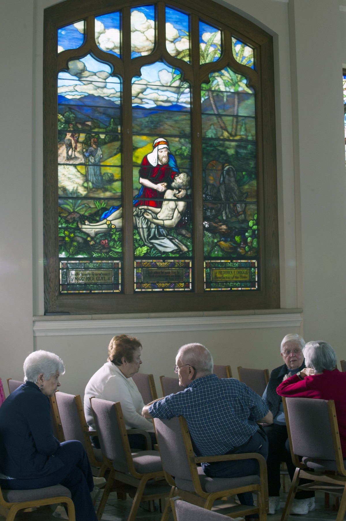 A group of people seated in a church, beneath a large stained glass window.