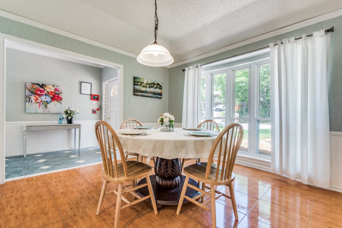 A cozy dining room featuring a round wooden table with chairs, set for a meal under a decorative chandelier, and brightened by natural light through large windows.