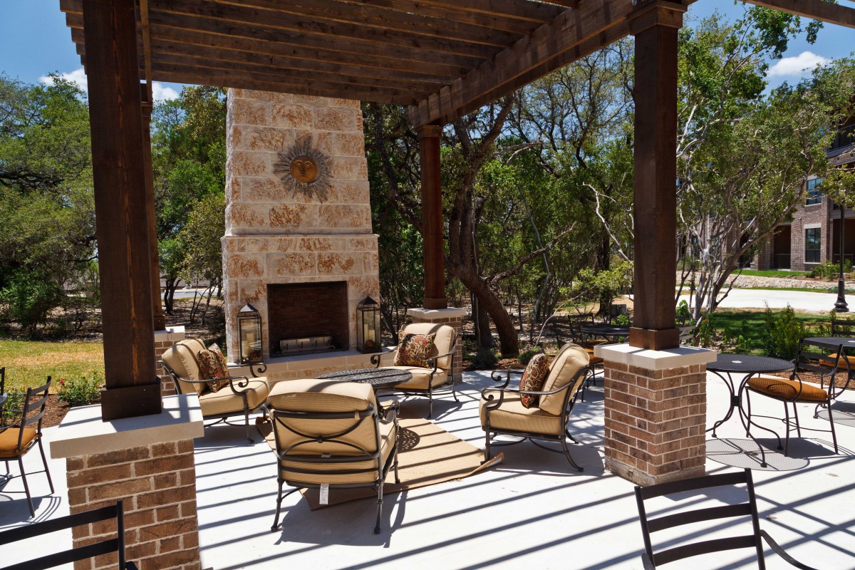 A cozy outdoor patio with a stone fireplace and comfortable seating under a wooden pergola, surrounded by trees.