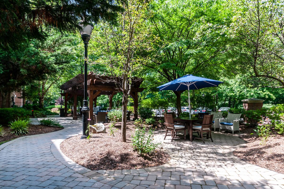 A peaceful garden setting with outdoor seating under a blue umbrella, surrounded by trees and a brick pathway.