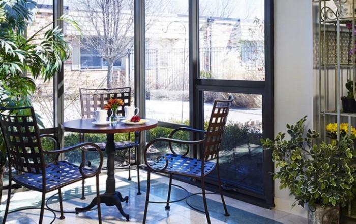 A cozy indoor dining area with a table, chairs, and sunlight streaming through the large windows.