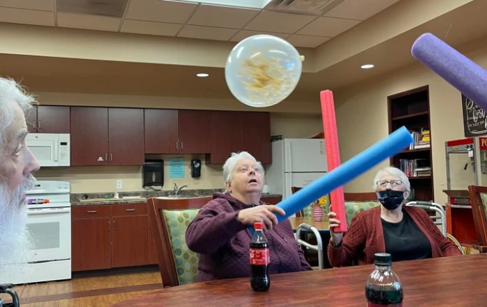 Elderly individuals enjoying a fun indoor game with balloons and pool noodles.