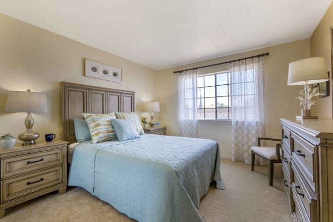 A cozy bedroom with a bed covered in light blue bedding, softly lit by a window draped with sheer curtains.