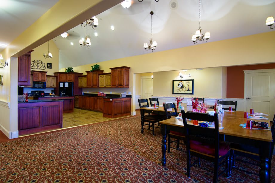 A spacious dining area with wooden furniture and a connected kitchen, featuring warm lighting and a carpeted floor.