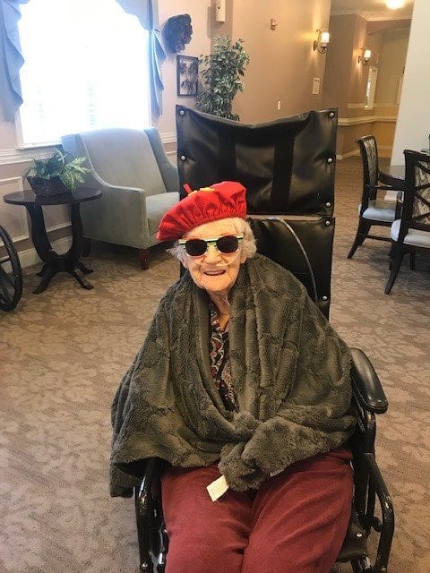 An elderly person sitting in a wheelchair indoors, wearing a red hat and sunglasses with a cheerful expression.