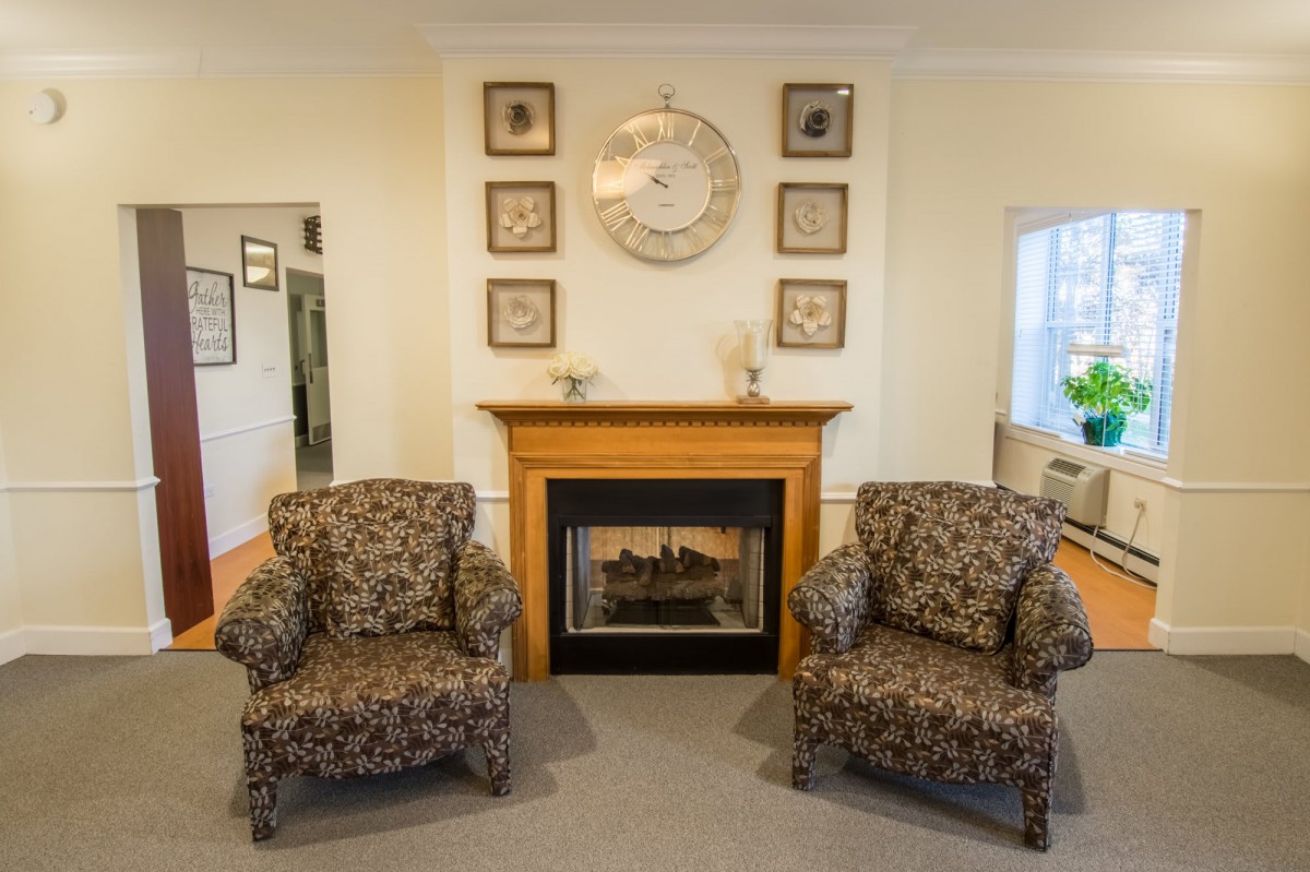 A cozy living room features a fireplace flanked by two patterned armchairs, with a decorative clock and wall art above.