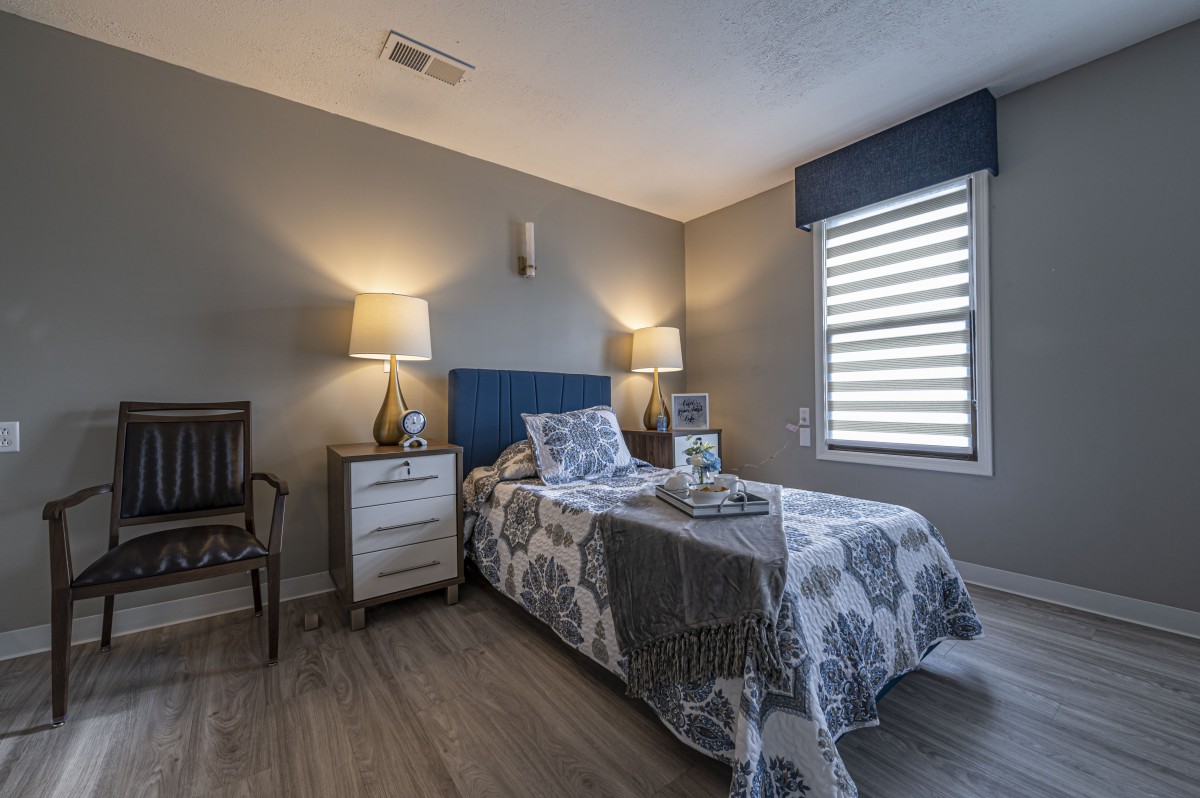 A modern bedroom featuring a single bed with floral bedding, flanked by two nightstands and lamps, under a window with blinds.