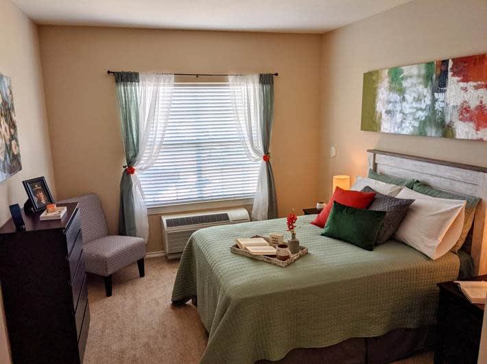 A cozy bedroom with neatly arranged decor, featuring a bed, window with curtains, and a comfortable chair.