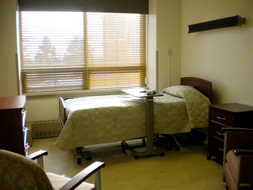 A well-lit hospital patient room featuring a bed, a chair, and a window with blinds.
