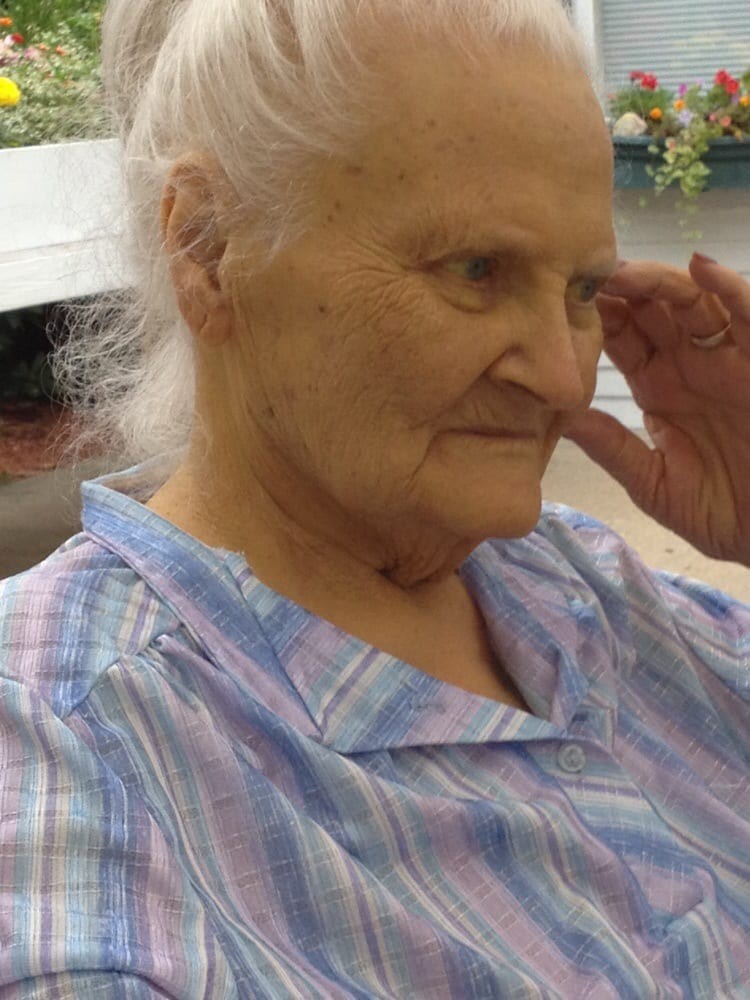 An elderly woman with gray hair is sitting indoors, wearing a plaid shirt.
