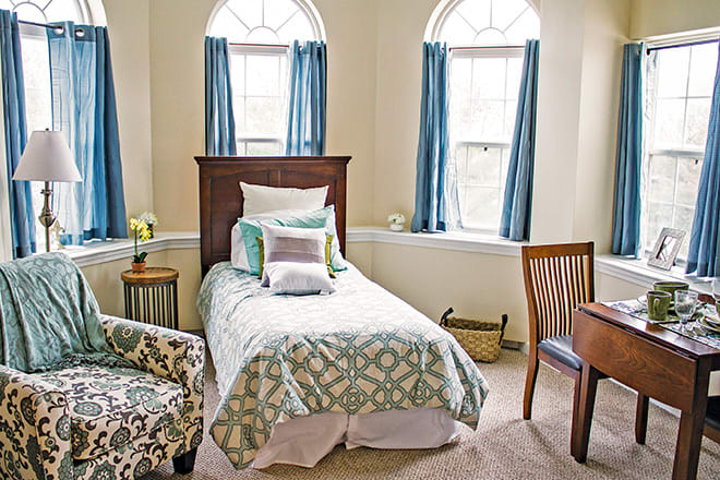 A cozy bedroom with a single bed, adorned with decorative pillows, next to large windows with blue curtains. An armchair and wooden desk complete the setup.