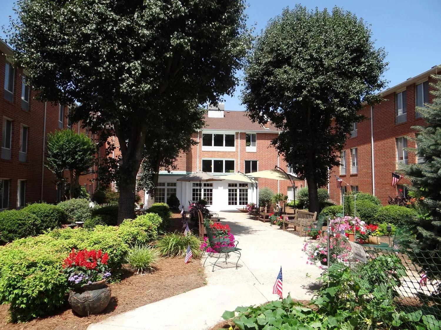 A serene garden courtyard with lush greenery, vibrant flowers, and outdoor seating leading to a multi-story brick building.