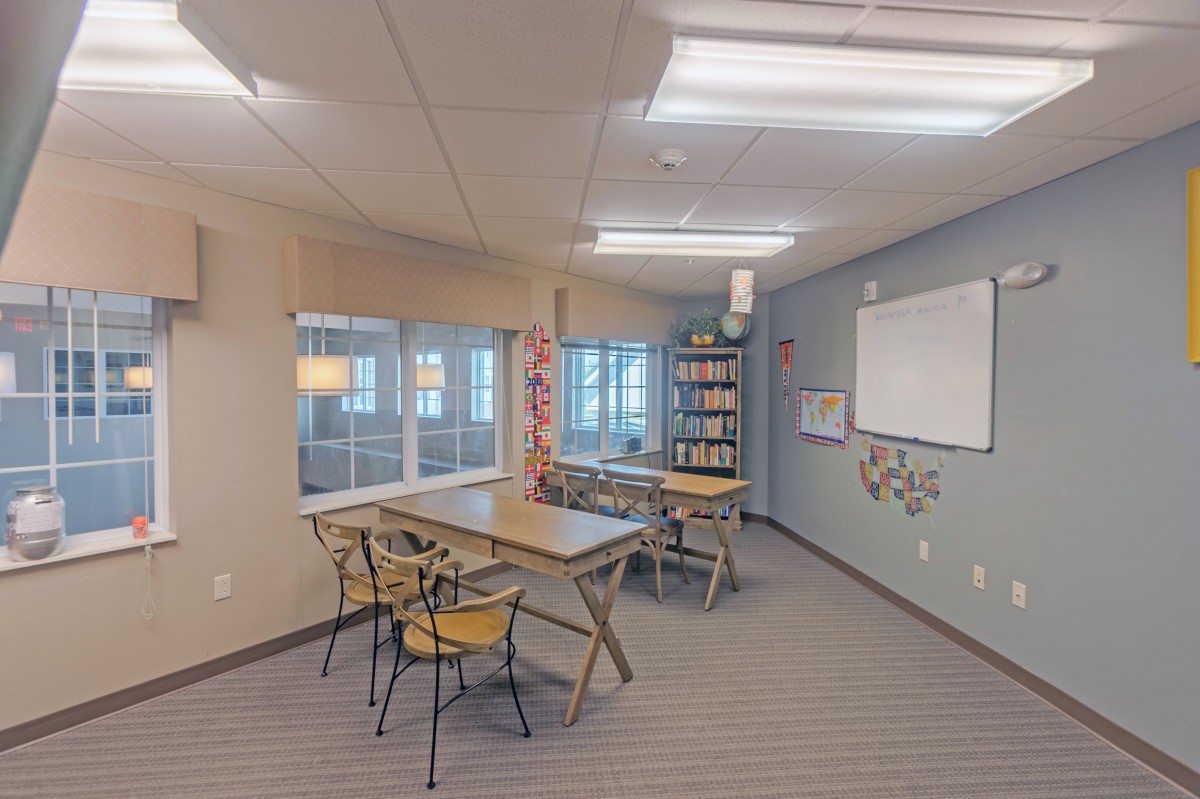 A cozy study area with tables, chairs, and a bookshelf next to large windows.