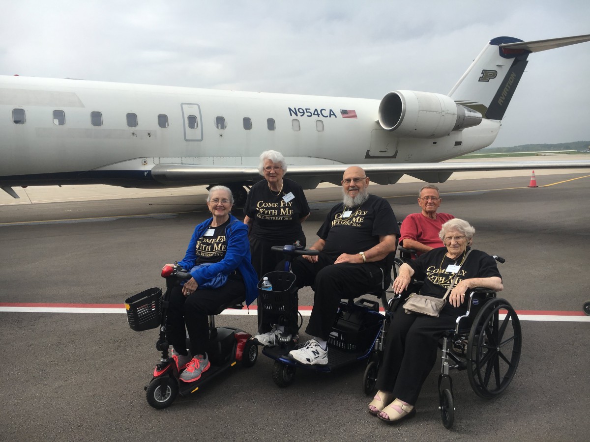 A group of elderly individuals, some in wheelchairs, posing near an airplane at the airport.