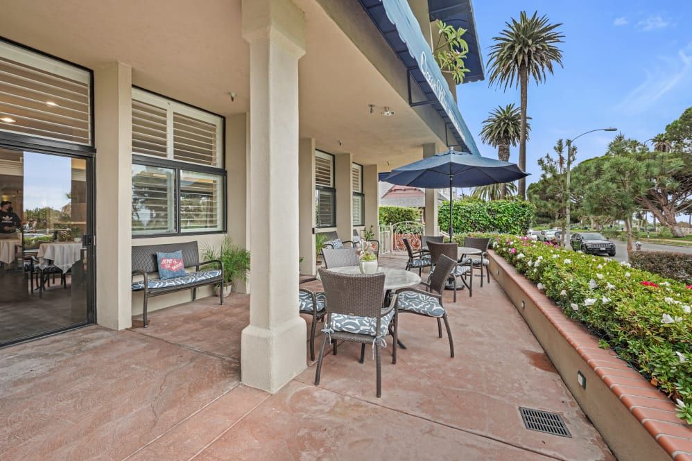 An outdoor patio area with tables and chairs under umbrellas, surrounded by lush greenery and palm trees.