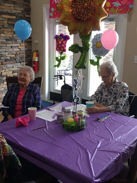Two elderly women sitting at a table during a celebration with balloons and decorations.