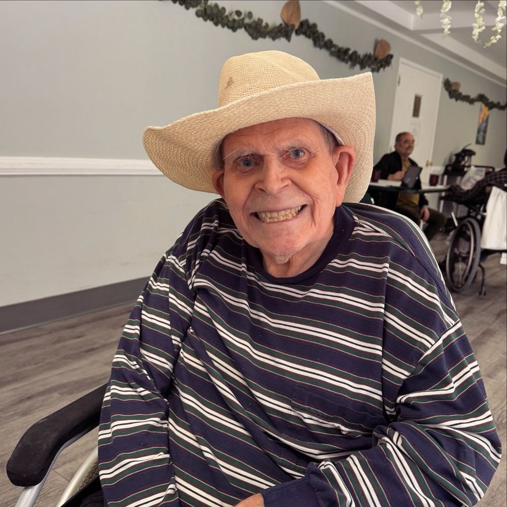 An elderly man wearing a cowboy hat and striped shirt, smiling while seated indoors.