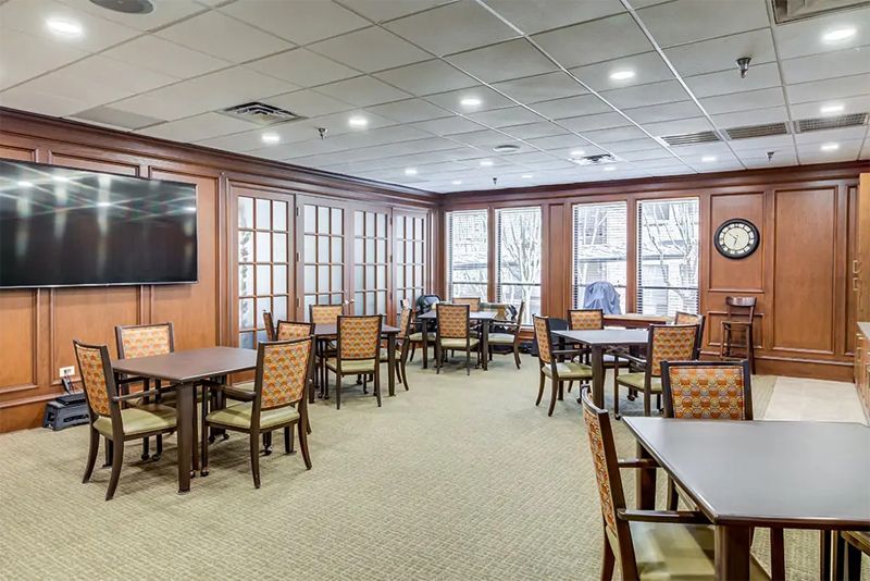 A well-lit dining room with multiple tables and chairs, featuring wood paneling and large windows.