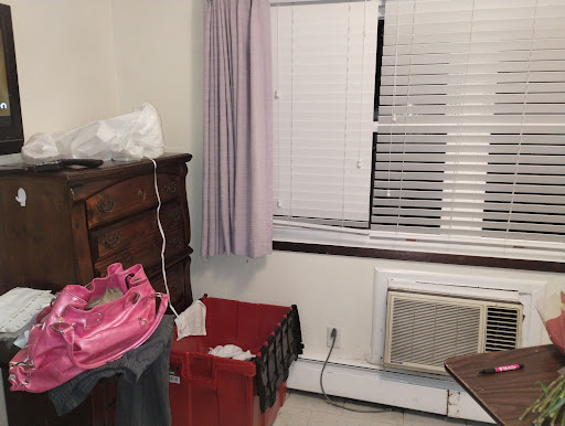 A cozy room featuring a dresser, window with curtains, and an air conditioner, alongside some clothing and storage baskets.