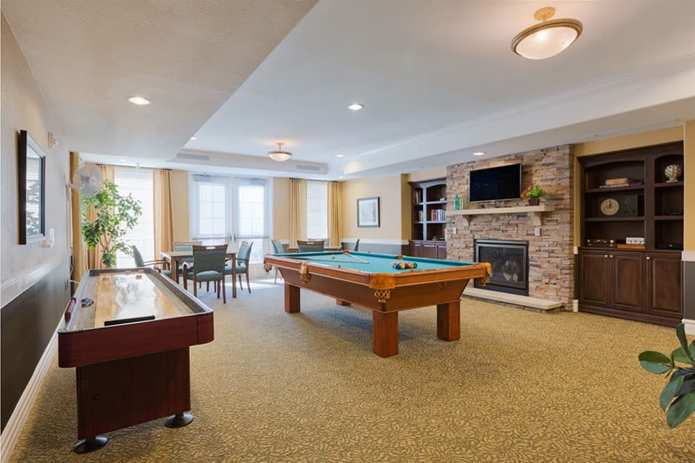 A cozy indoor game room featuring a pool table, shuffleboard, and a fireplace surrounded by a seating area.