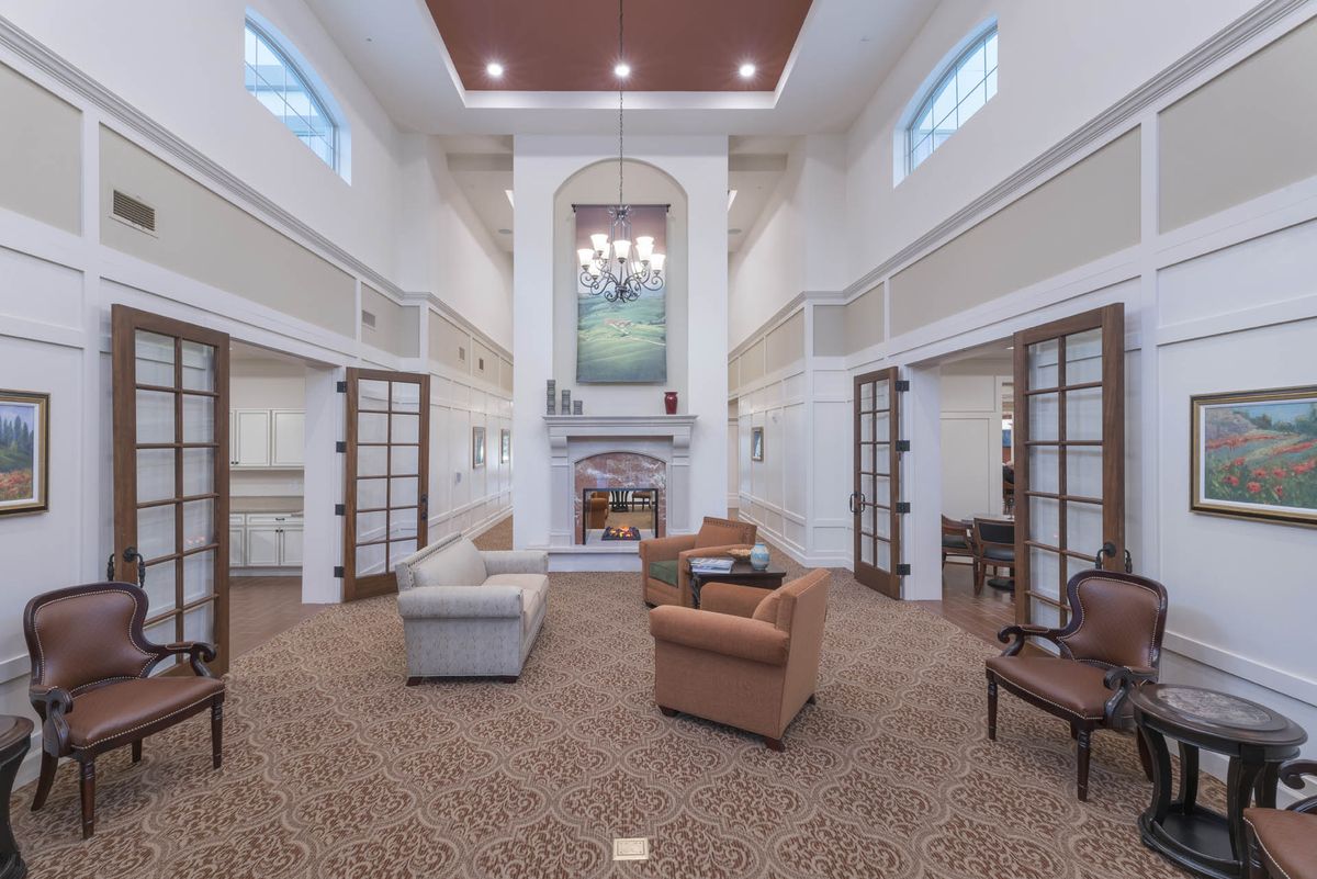 A spacious living room with a high ceiling, featuring a seating area around a central fireplace and elegant chandeliers.