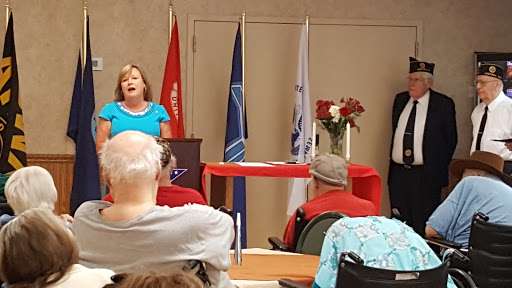 A speaker delivers a presentation at a community event, with an audience gathered and various flags displayed.
