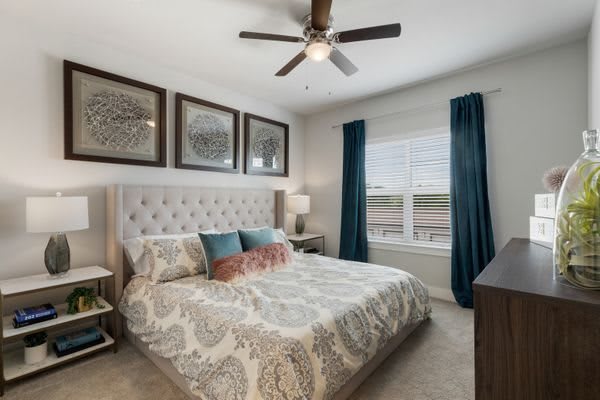 A cozy bedroom with a tufted headboard, patterned bedding, and blue curtains decorating the window.
