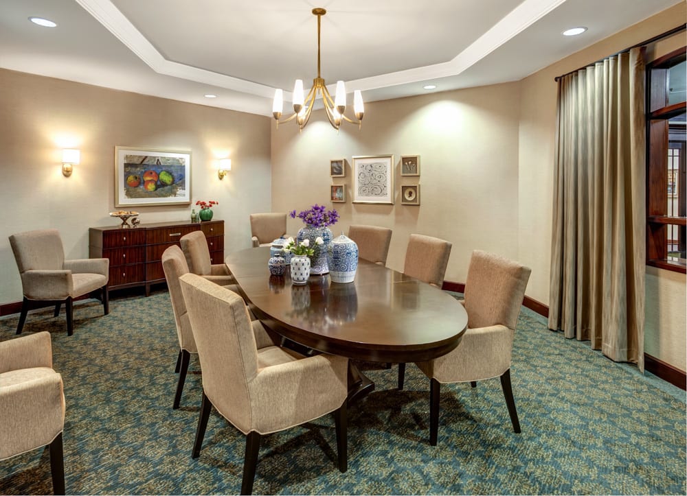 An elegant dining room featuring a large oval table surrounded by upholstered chairs, with tasteful artwork adorning the walls and a chandelier providing soft lighting.