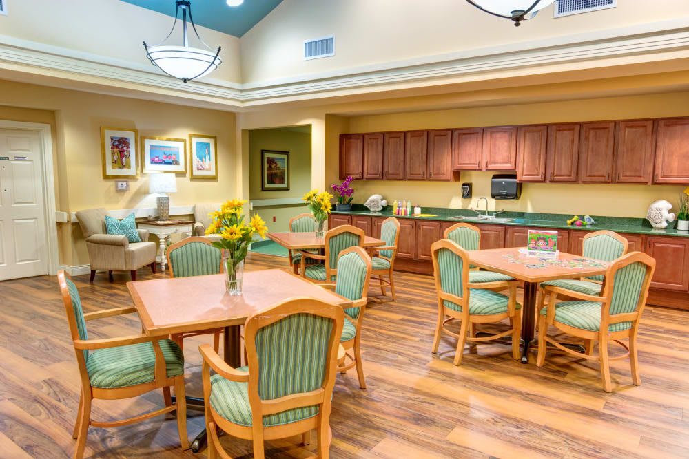 A brightly lit dining area with wooden tables and green cushioned chairs, featuring sunflowers as centerpieces and a kitchenette in the background.