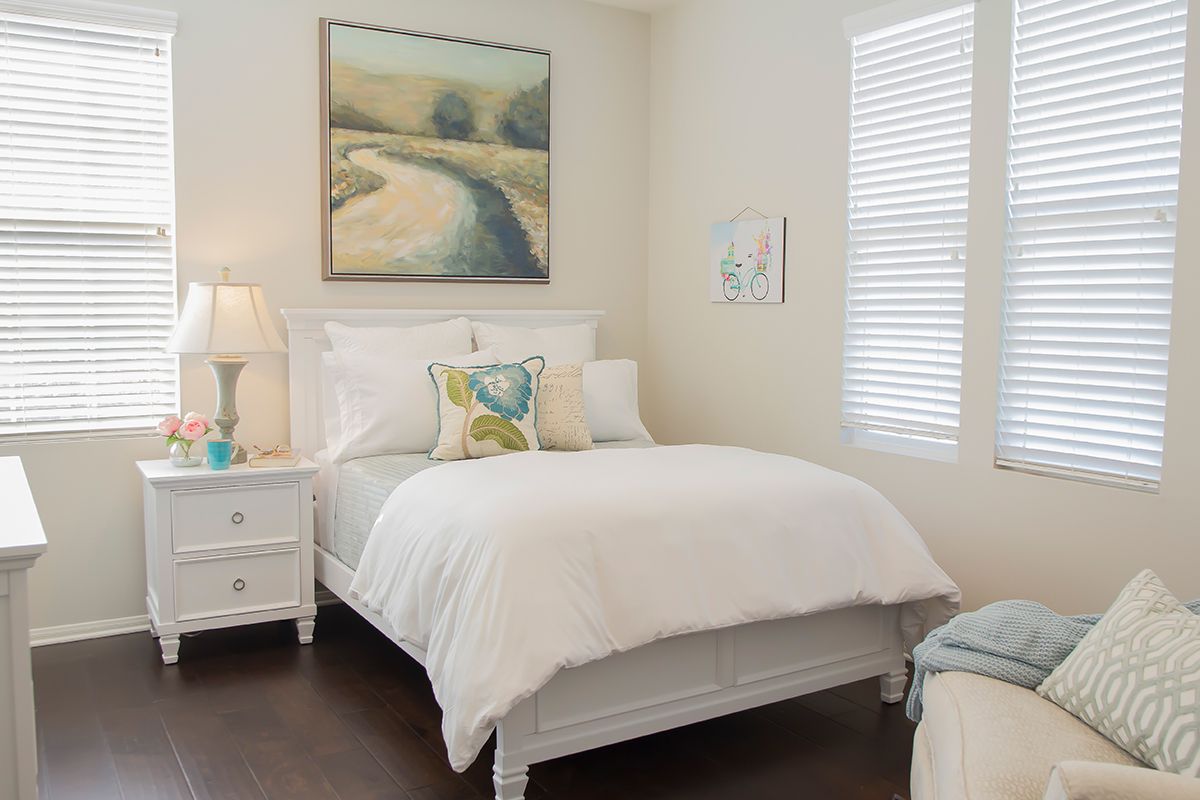 A cozy bedroom featuring a white bed with decorative pillows, a nightstand with a lamp, and artwork on the walls.