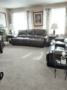 A cozy living room featuring dark leather sofas, soft carpeting, and natural light from large windows.