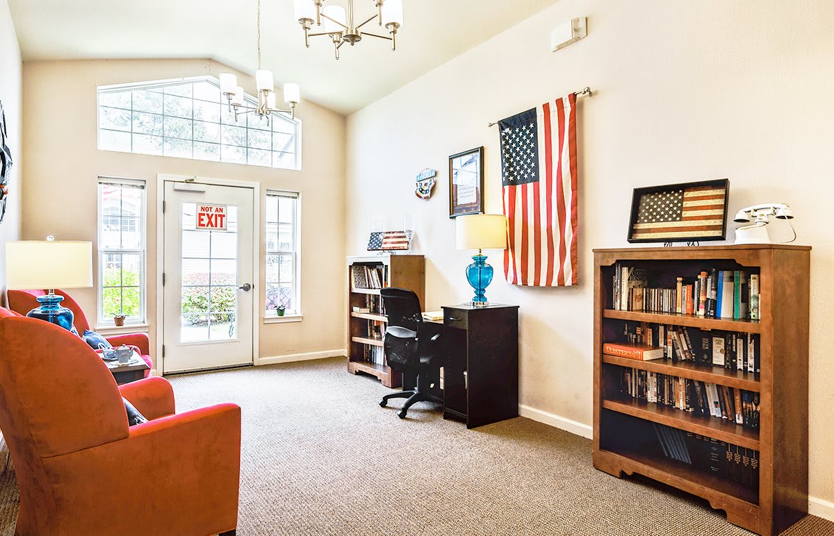 A brightly lit office interior featuring bookshelves, a desk with a chair and lamp, and an American flag on the wall.