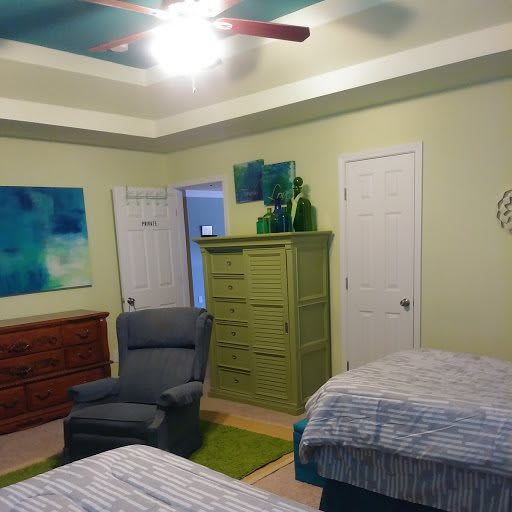 A cozy bedroom with a chest of drawers, armchair, and twin beds. The room is decorated with artwork and features a ceiling fan.