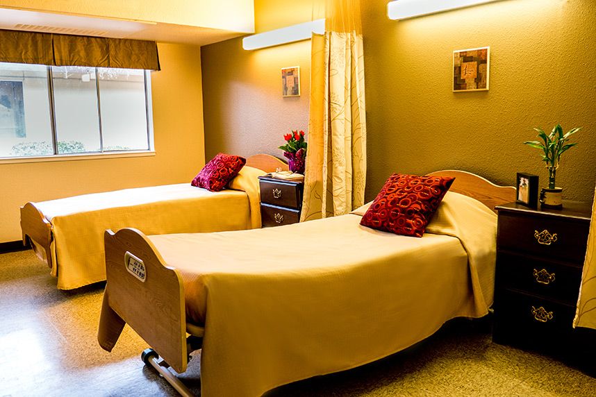 A brightly lit nursing home or hospital room with two beds covered in yellow bedspreads and red pillows.