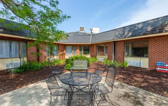 A tranquil courtyard with a metal outdoor table and chairs surrounded by greenery and a brick building.