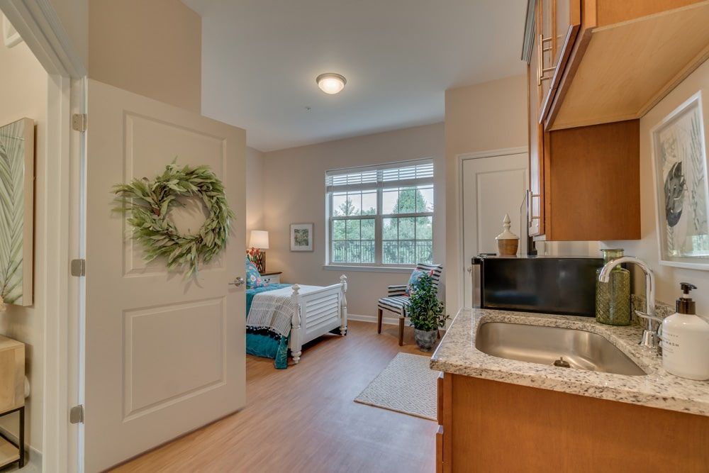A cozy bedroom with a kitchenette, featuring natural light and decorative touches like plants and wall art.
