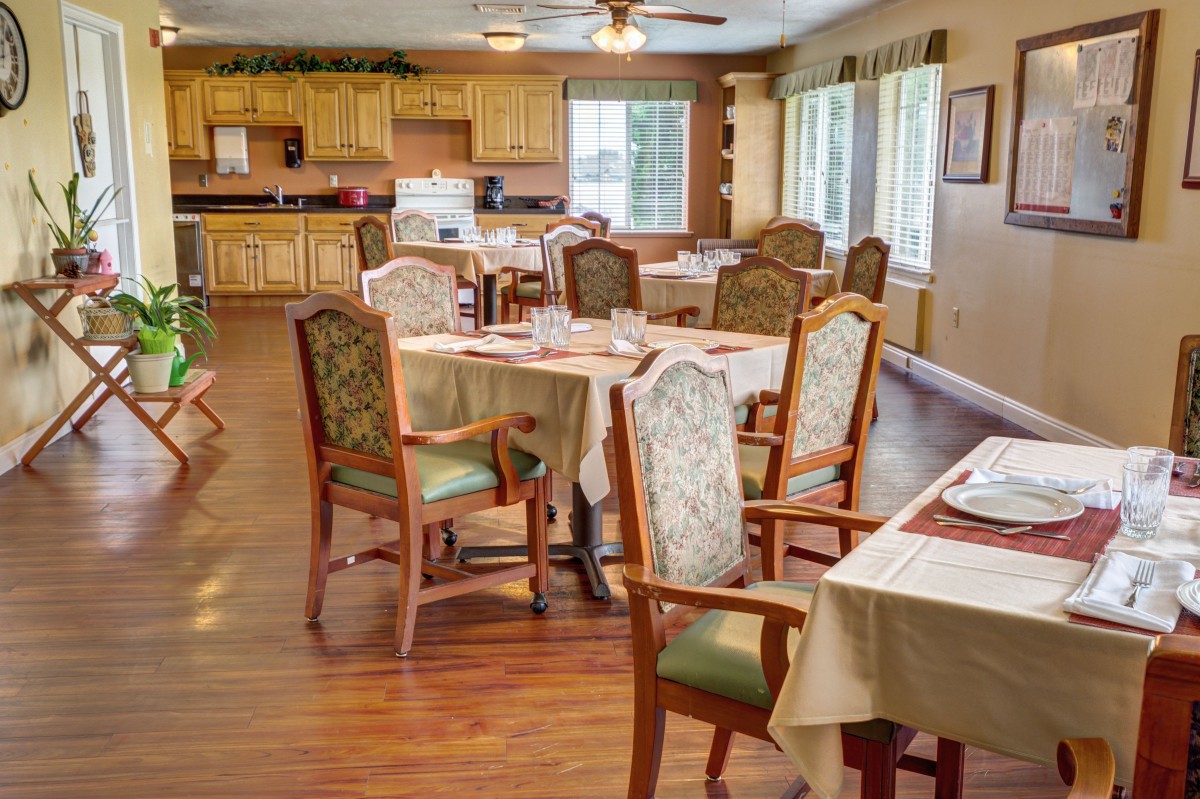 A cozy dining room with neatly arranged tables and chairs, featuring wooden flooring and large windows with curtains.