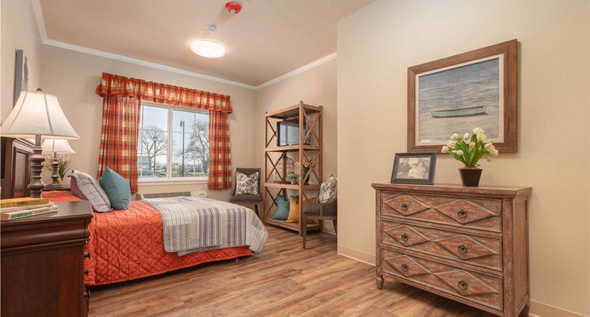 A cozy bedroom with wooden flooring, featuring a neatly made bed, a dresser with artwork on top, and a shelf by the window.
