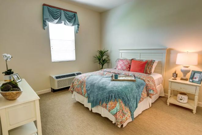 A cozy bedroom with a neatly made bed, colorful pillows and blankets, a window allowing natural light, and a plant in the corner.