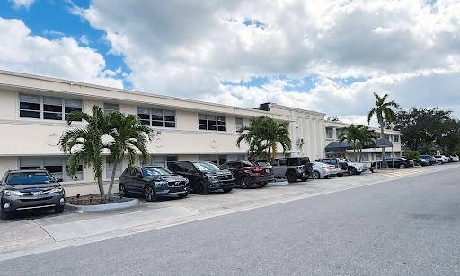 A row of parked vehicles along a building, framed by palm trees under a partly cloudy sky.